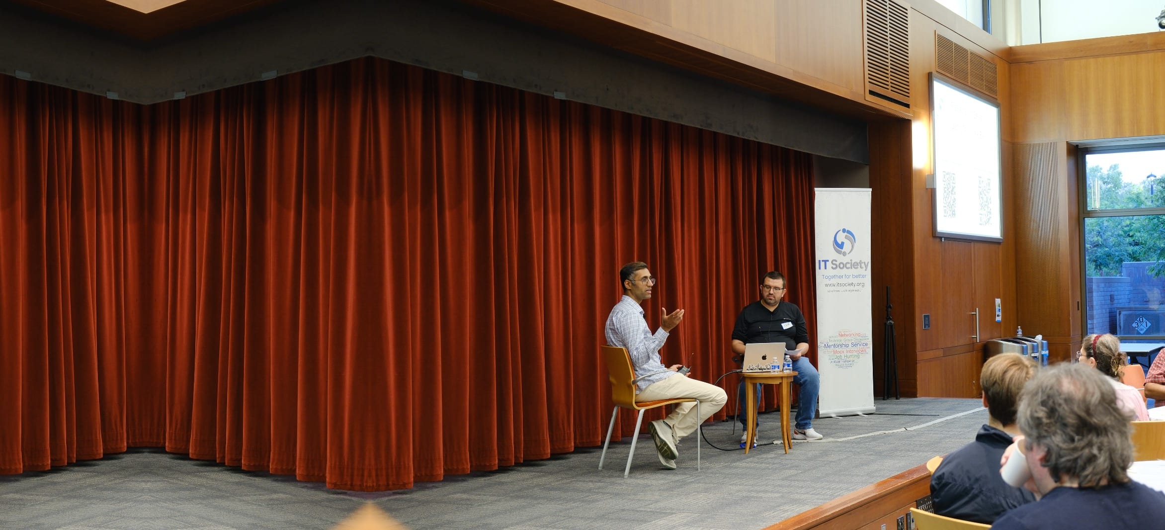 Two speakers in conversation on stage beside the IT Society banner